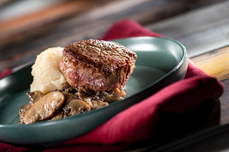 A beautifully presented filet mignon served on a green plate alongside mashed potatoes and sautéed mushrooms. The dish is arranged with an elegant presentation on a textured wooden surface, suggesting a gourmet dining experience.
