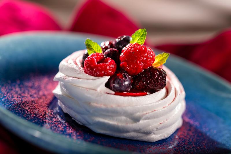 The image showcases a beautifully plated pavlova dessert, featuring a delicate meringue shell topped with a vibrant assortment of fresh berries and mint leaves. The dessert is served on a blue plate, with a softly blurred red fabric in the background.
