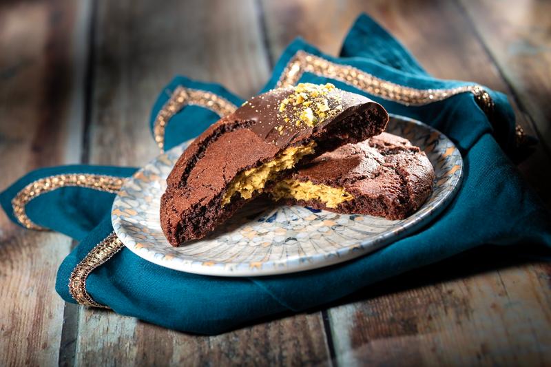 A close-up image of a split cookie, showcasing a chocolate exterior and a creamy, pistachio filling. The cookie sits on a decorative plate, placed atop an elegant teal fabric.