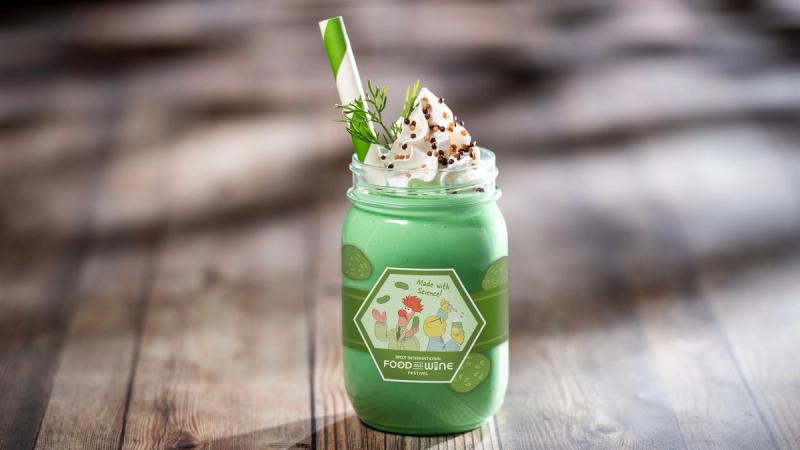 This image features a vibrant green milkshake served in a mason jar, topped with whipped cream and garnished with herbs. The drink is styled with a green and white striped straw, resting on a wooden surface with natural lighting.