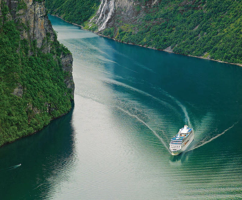 A cruise ship navigates through a serene fjord, surrounded by lush green cliffs and tranquil waters. The landscape showcases natural beauty, characterized by steep mountains and clear blue water.