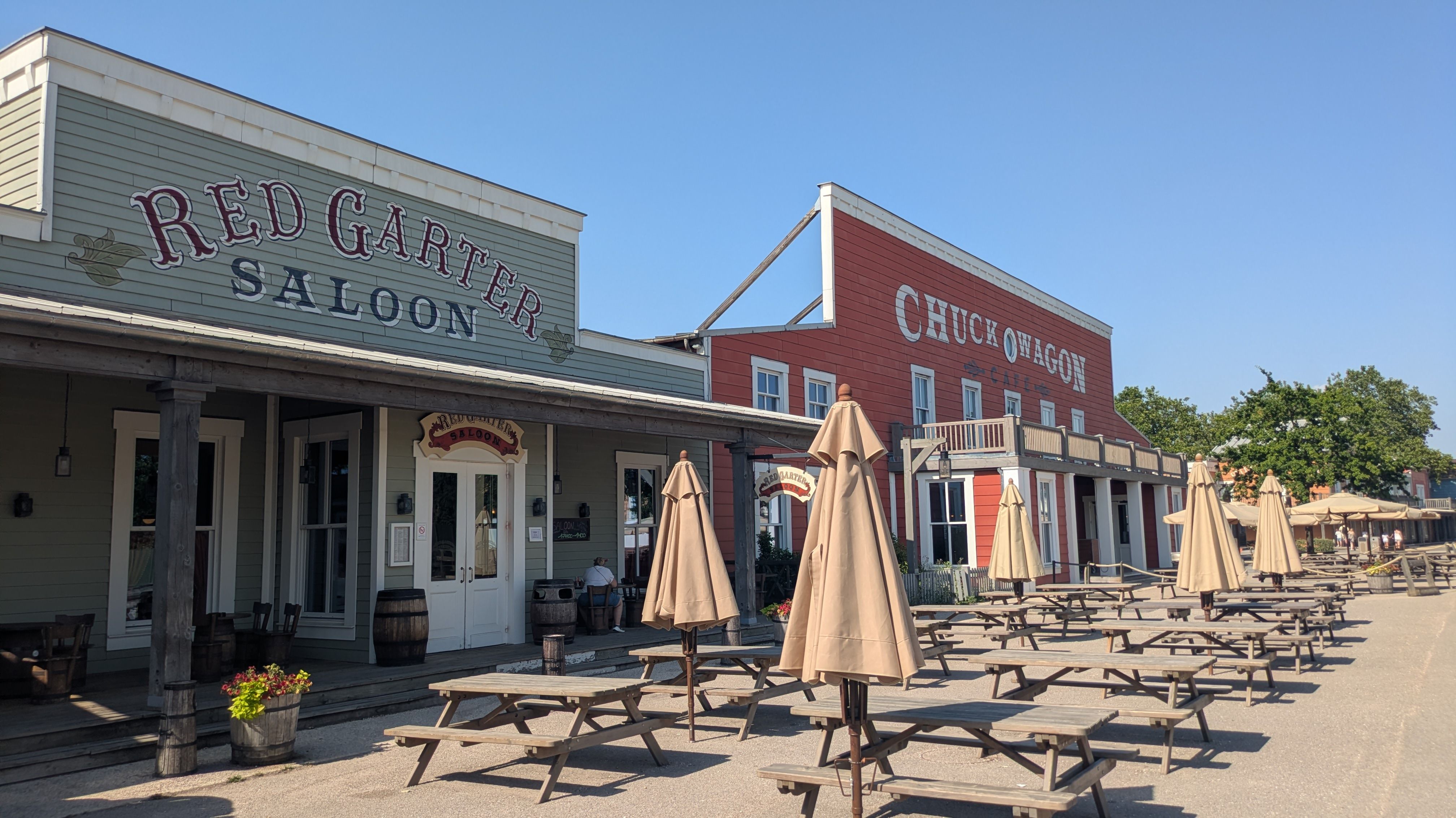 A view of a Western-style town featuring two saloons, 'Red Garter Saloon' and 'Chuckwagon,' with empty outdoor seating areas and umbrellas. The scene is set under a clear blue sky, showcasing a rustic, old-time ambiance.