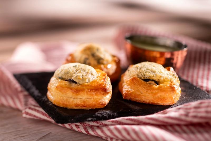 The image displays a close-up view of three golden-brown pastry bites filled with an unknown creamy substance. They are arranged on a dark slate plate, accompanied by a small bowl of green sauce, with a red and white striped cloth underneath.