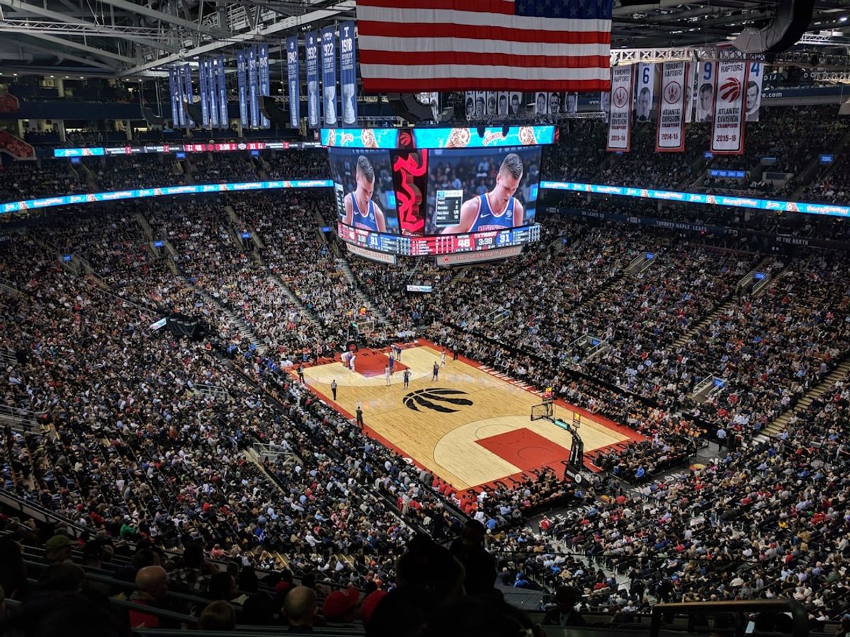 A large indoor basketball arena filled with fans watching a game. The scoreboard displays player statistics and the game's progress, while the court features the team's logo at center court.