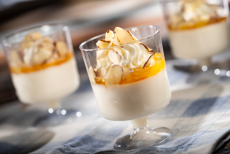 The image features three glasses of panna cotta, a classic Italian dessert, topped with a layer of orange sauce and garnished with almond slices. The dessert is arranged on a textured tablecloth, with soft lighting enhancing its creamy texture.