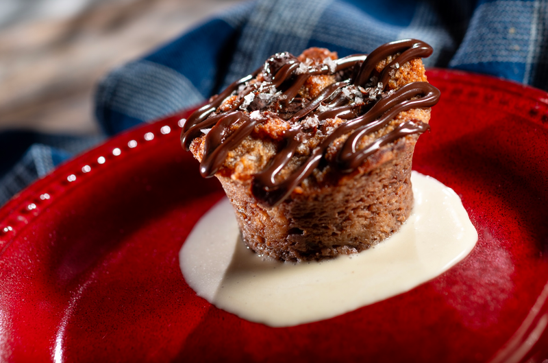 The image showcases a freshly made bread pudding served on a red plate. The dessert is topped with chocolate drizzle and accompanied by a creamy sauce, highlighting its indulgent nature.
