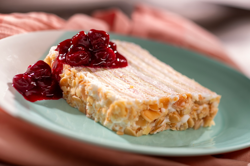 The image features a slice of a dessert layered with cream and nuts, garnished with a red fruit compote on the side. The dessert is presented on a light blue plate against a soft, blurred background.