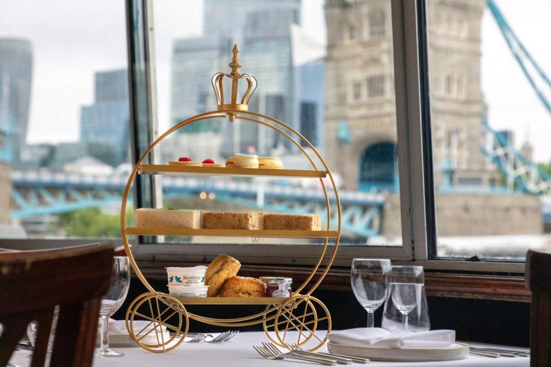 A luxurious afternoon tea setup featuring a gold carriage-shaped stand with pastries, cakes, and sandwiches. The table is set with elegant glassware and is positioned near large windows offering a view of London’s Tower Bridge and modern skyscrapers.
