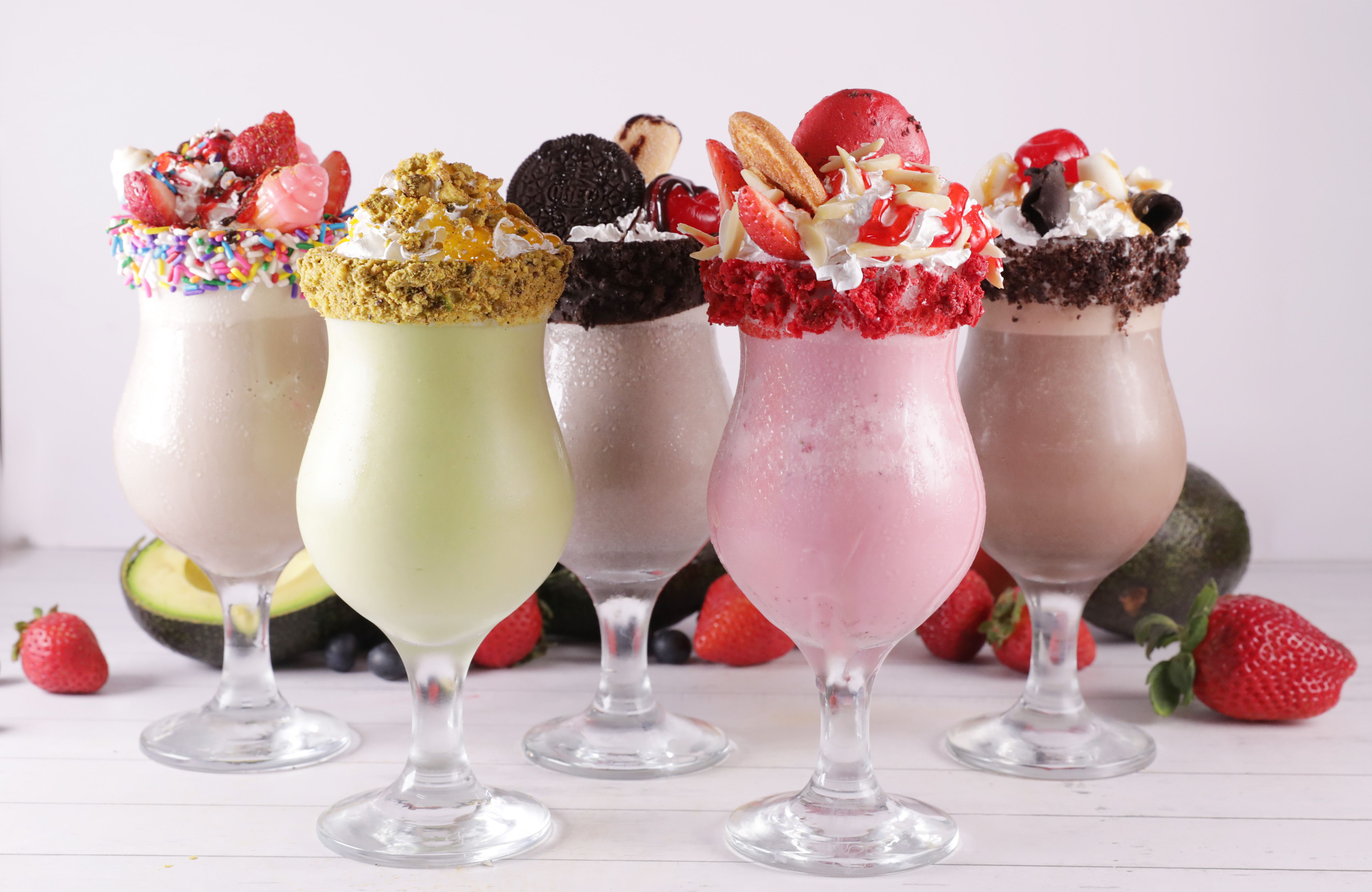 A colorful array of five milkshakes in decorative glasses, each topped with various garnishes such as fruits, whipped cream, and cookies. The milkshakes come in different flavors and colors, highlighting a vibrant and enticing display.