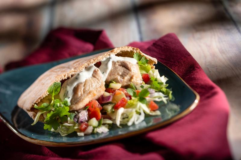 The image features a close-up of a pita filled with chicken, lettuce, tomatoes, and a creamy sauce, presented on a teal plate. The background includes a textured wooden surface and a soft fabric napkin, enhancing the culinary appeal of the dish.