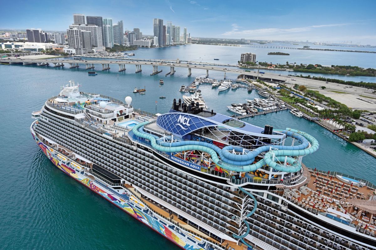 The image depicts a Norwegian Cruise Line ship docked in Miami, with its colorful exterior and a water slide on top. The background features the Miami skyline and a marina filled with boats.