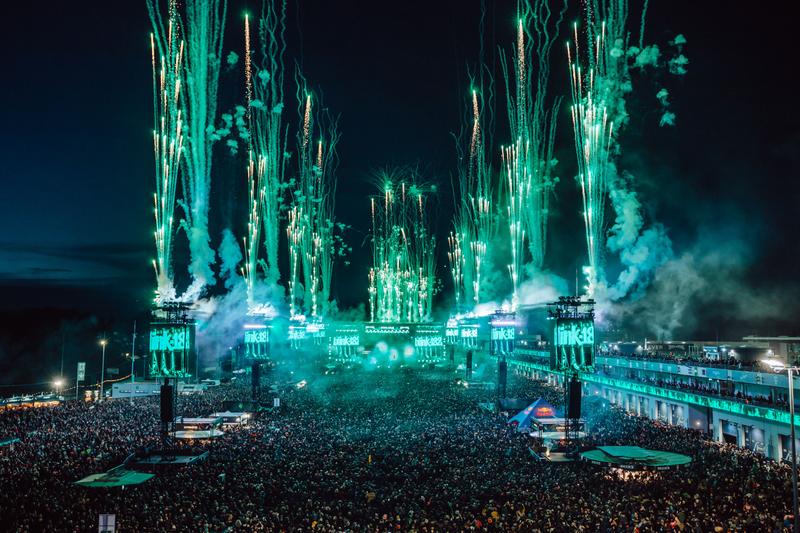 A large crowd gathers at an outdoor music festival, with vibrant fireworks lighting up the night sky. The stage is prominently featured, surrounded by thousands of spectators enjoying the performance.