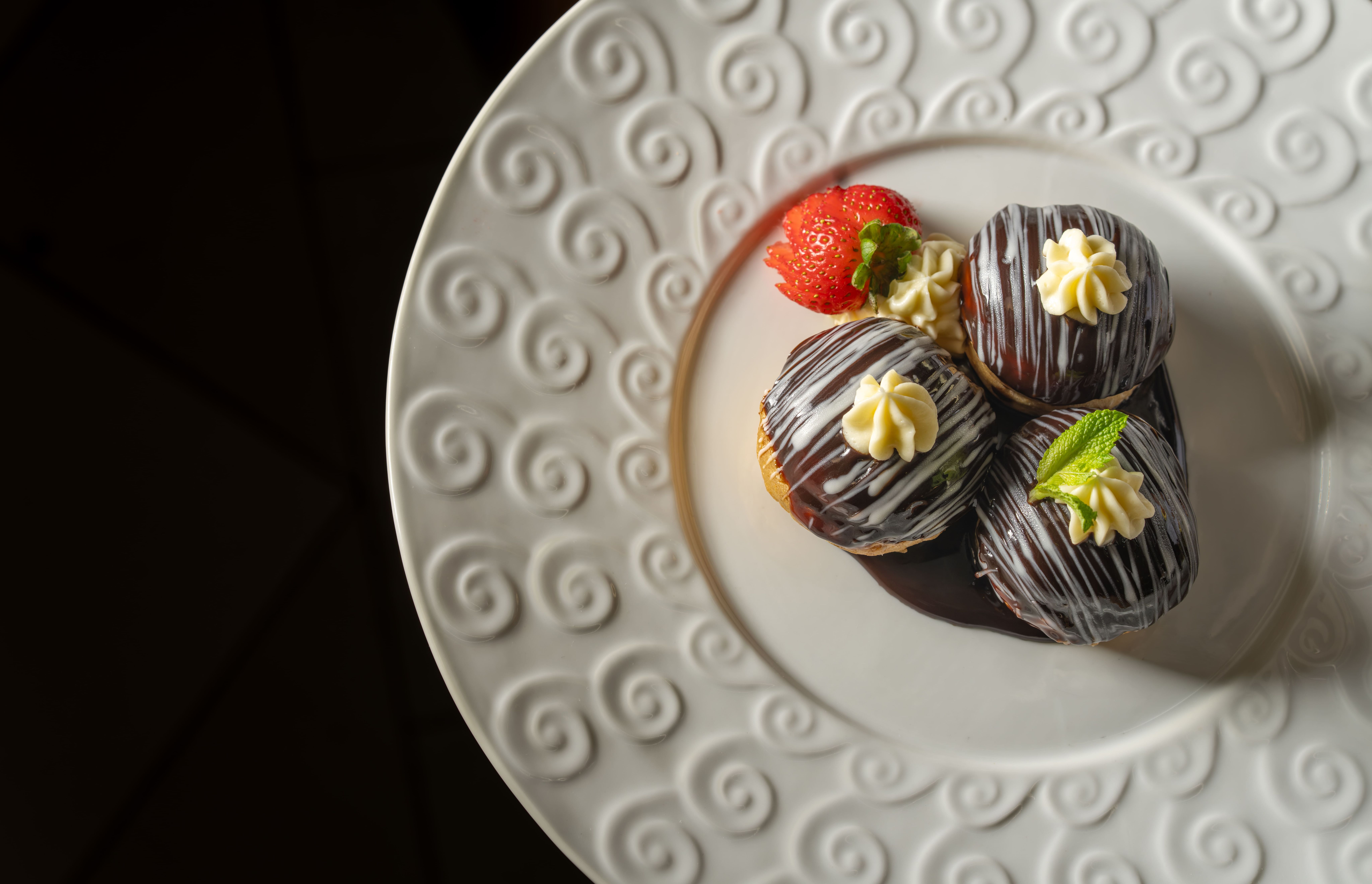 The image features a close-up of three chocolate-covered pastries arranged on an elegant decorative plate. A fresh strawberry and a mint leaf garnish complement the desserts, creating an appealing visual presentation.