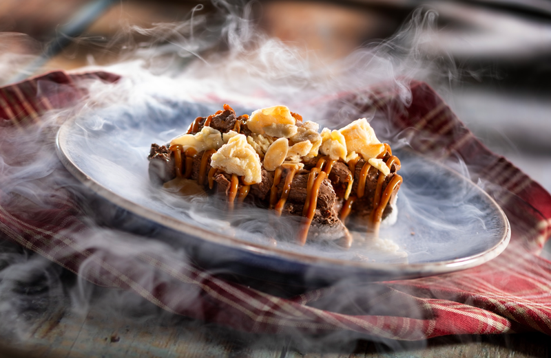 The image features a gourmet dessert showcasing a truffle mousse topped with various ingredients like pretzels, nuts, and a drizzle of caramel. The dish is presented on a plate and surrounded by a delicate cloud of smoke, suggesting a dramatic culinary experience.