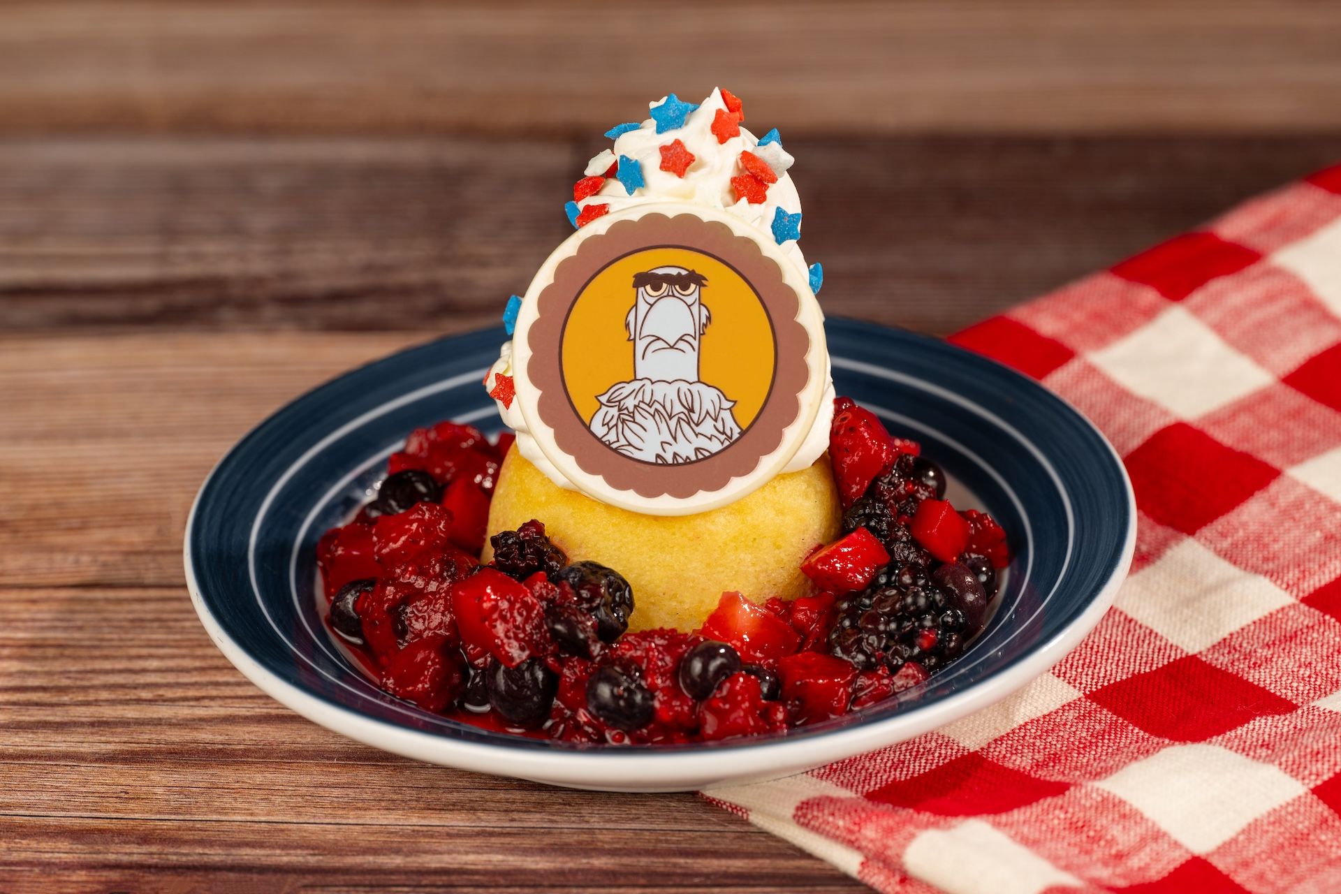 The image features a dessert composed of a yellow cake topped with whipped cream and a circular decoration featuring a cartoon eagle. The cake is surrounded by a colorful mix of berries including strawberries and blueberries, all presented on a patterned tablecloth.