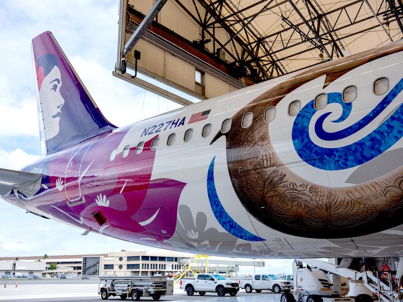 The image shows a close-up view of a colorful airplane with Hawaiian-themed graphics, parked inside a hangar. The design features intricate patterns representative of Hawaiian culture on the fuselage.