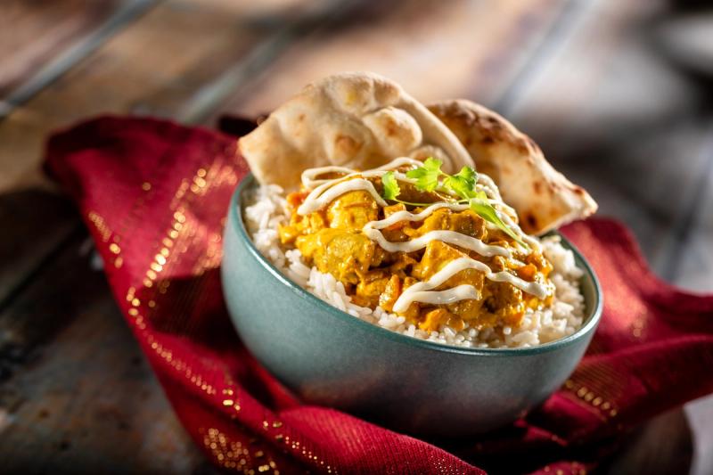 A bowl of chicken tikka masala served over a bed of rice, garnished with herbs and a drizzle of sauce. The dish is complemented by pieces of naan bread placed beside it on a vibrant cloth.