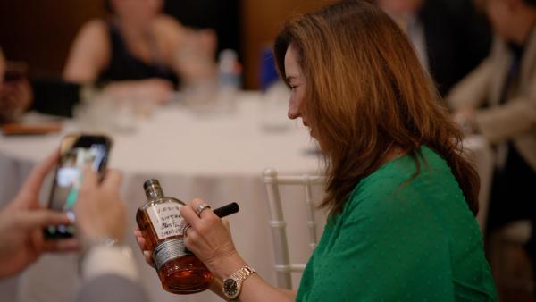 A woman is holding a bottle of whiskey while signing it for a fan at an event. The setting appears to be indoors with a blurred background of attendees.
