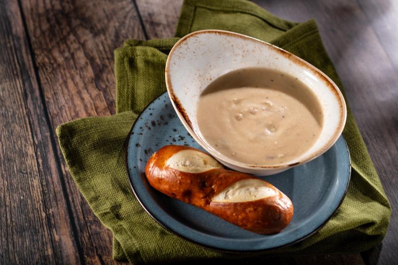 The image features a bowl of soup served with a twisted pretzel on a plate. The soup appears creamy and is set against a rustic wooden backdrop, creating a warm and inviting presentation.