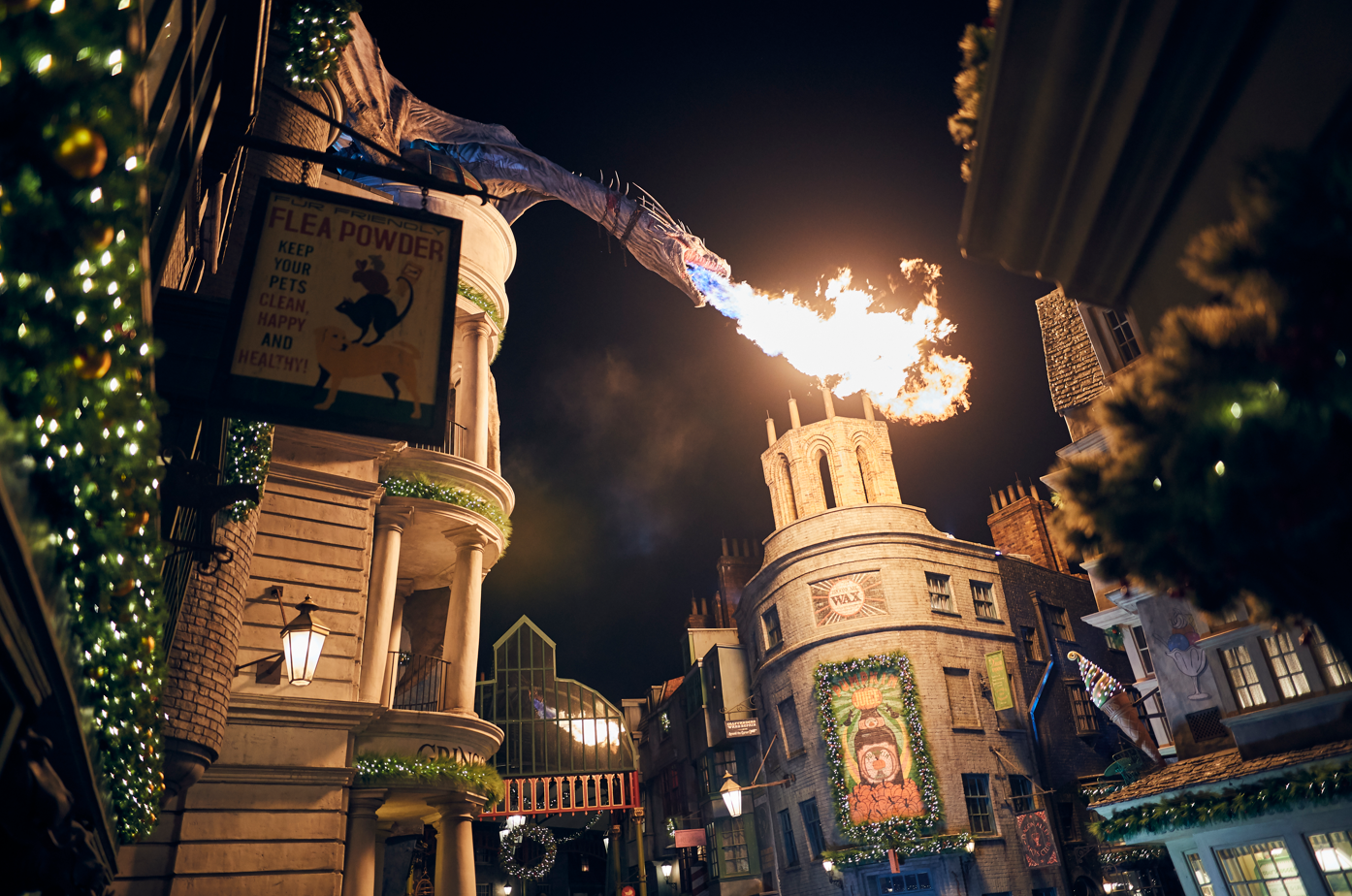 The image captures a night scene in a themed street resembling a magical marketplace. A dragon is prominently spewing fire over the buildings, creating a whimsical and vibrant atmosphere.