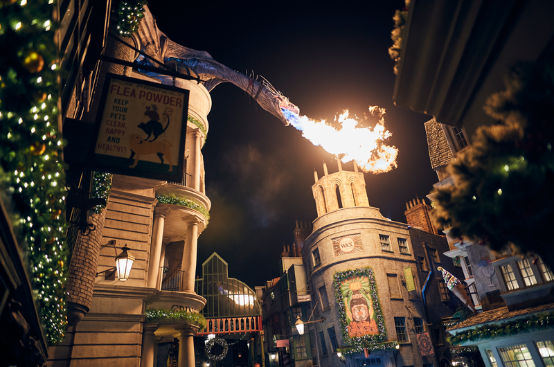 The image captures a night scene in a themed street resembling a magical marketplace. A dragon is prominently spewing fire over the buildings, creating a whimsical and vibrant atmosphere.