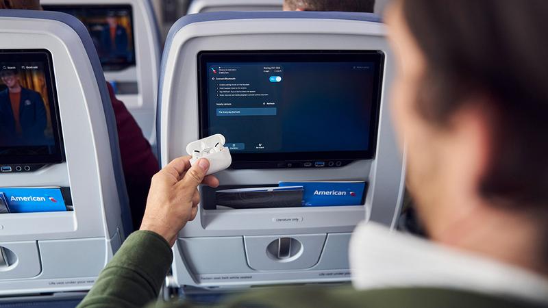 An individual is holding a pair of wireless earbuds while seated on an airplane. The seatbacks in front feature personal entertainment screens, displaying options for use during the flight.