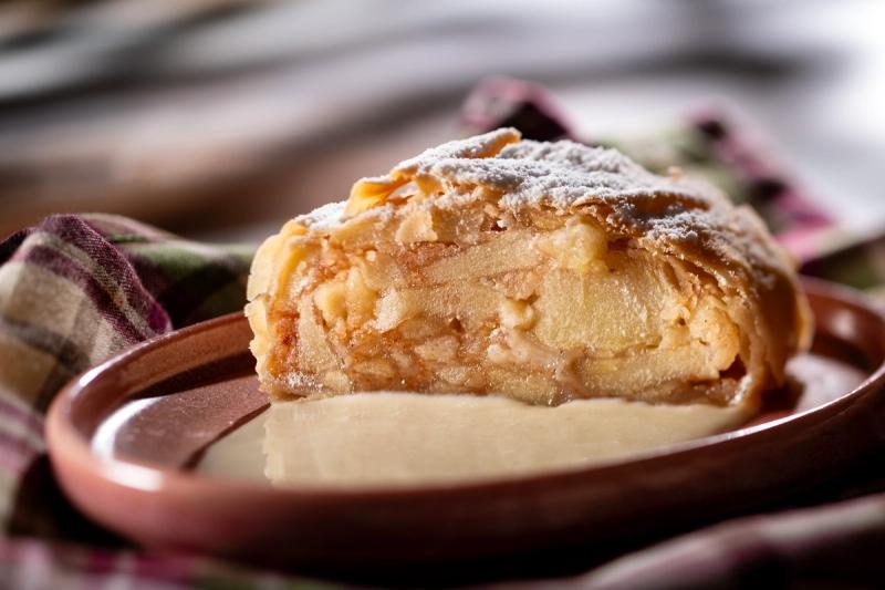 The image features a close-up of a slice of apple strudel on a plate, accompanied by a light sauce. The dessert appears freshly made, showcasing layers of pastry and visible apple filling.