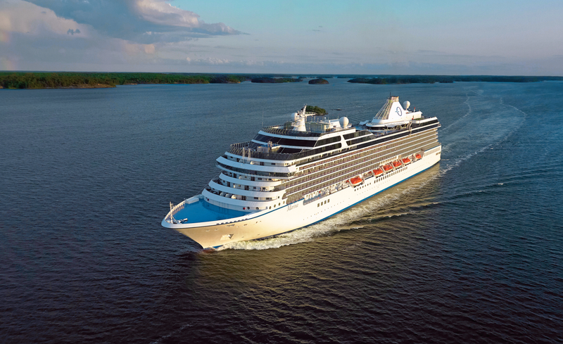 A large cruise ship sails through calm waters, surrounded by lush greenery and an expansive sky. The vessel showcases modern design features, with ample deck space and passenger amenities visible.