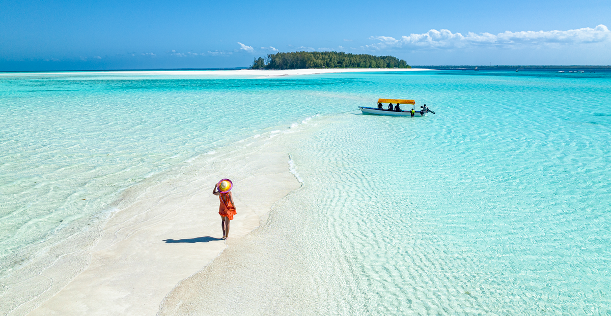 A person wearing an orange dress and a hat stands on a sandbar surrounded by clear turquoise waters. In the background, a small boat is visible near a lush green island under a bright blue sky.