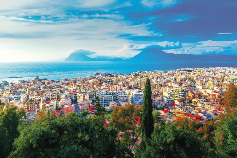 A picturesque view of a coastal city featuring vibrant buildings and lush greenery in the foreground. The background displays a tranquil sea and distant mountains, showcasing a serene landscape.