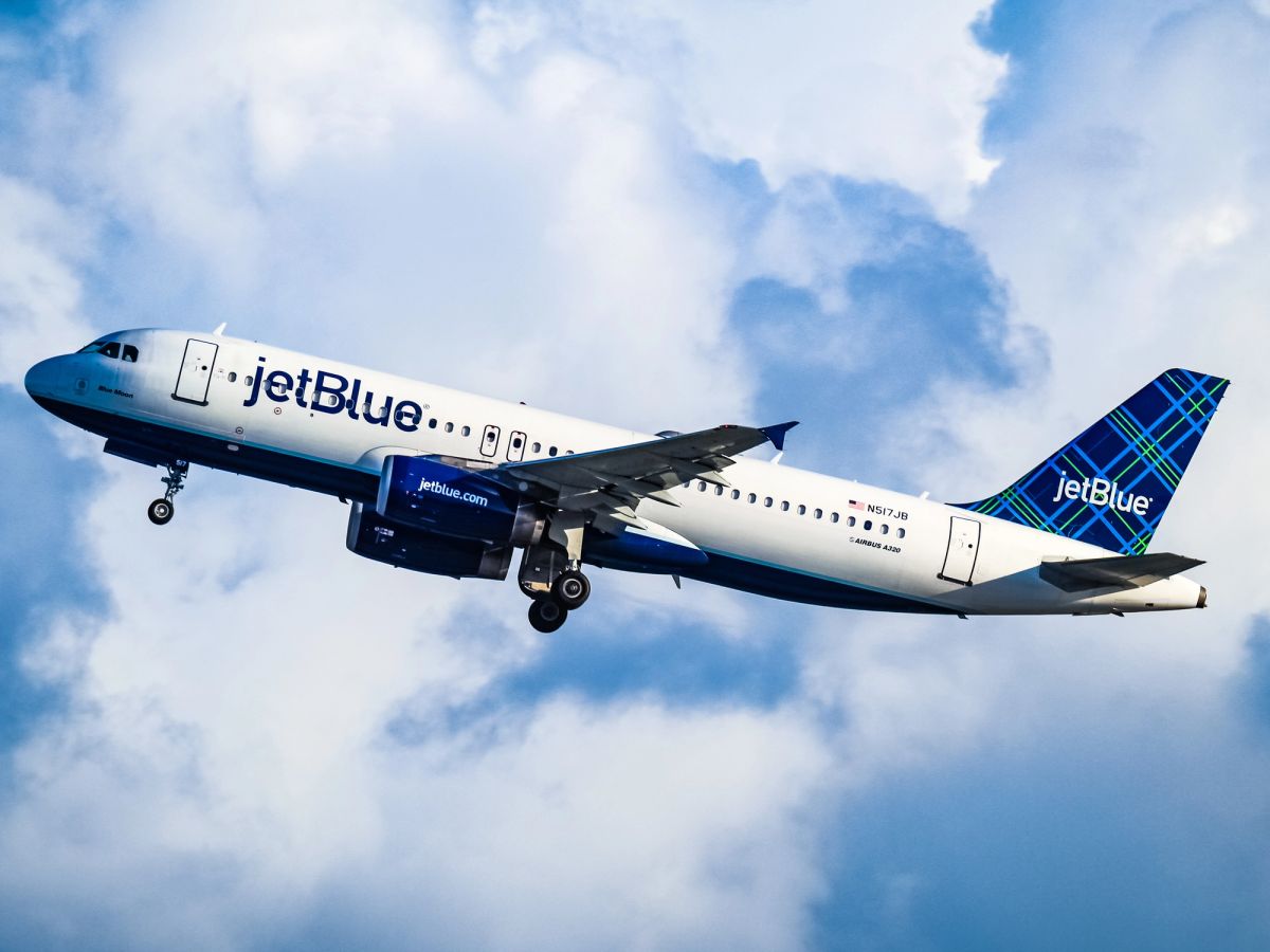 A JetBlue Airways airplane is captured in mid-flight against a backdrop of cloudy skies. The aircraft showcases the airline's distinctive blue and green plaid tail design.