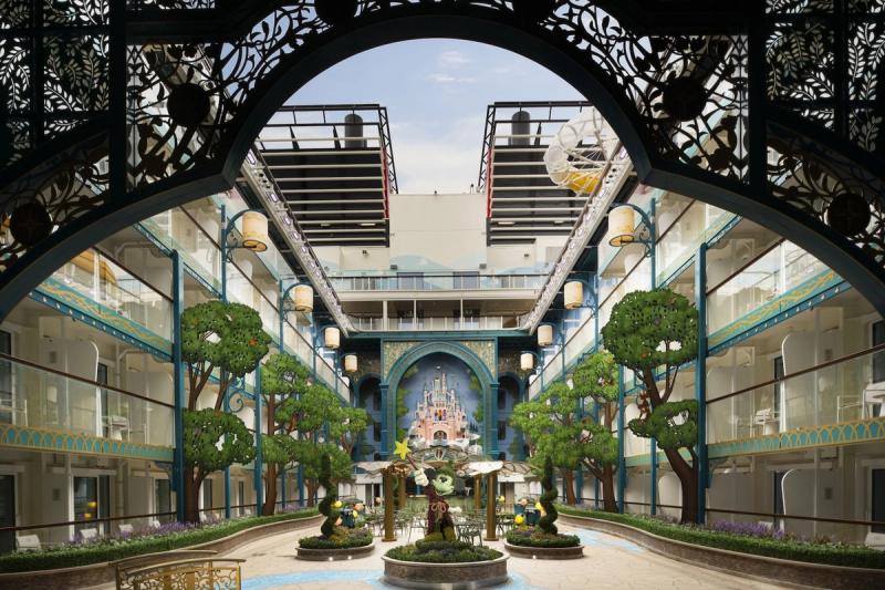 The image depicts an ornate courtyard of a hotel featuring greenery, a central fountain, and intricate architectural details. The design incorporates elements reminiscent of fantasy and playfulness, providing a vibrant and inviting atmosphere.