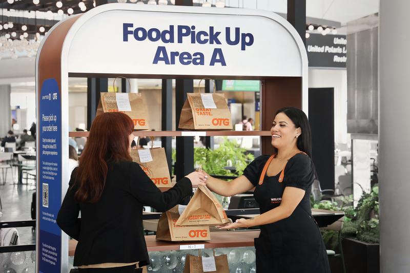 The image shows a food pickup area at an airport where a customer is receiving a takeout order from a staff member. The setting is bright and modern, indicating a focus on convenience for travelers.