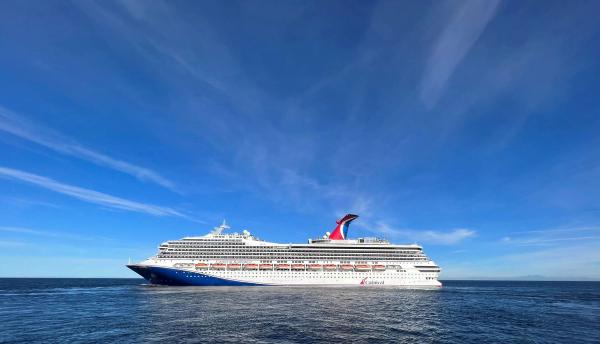 A large cruise ship sailing through calm waters under a clear blue sky. The ship features a prominent red and blue design along its hull.