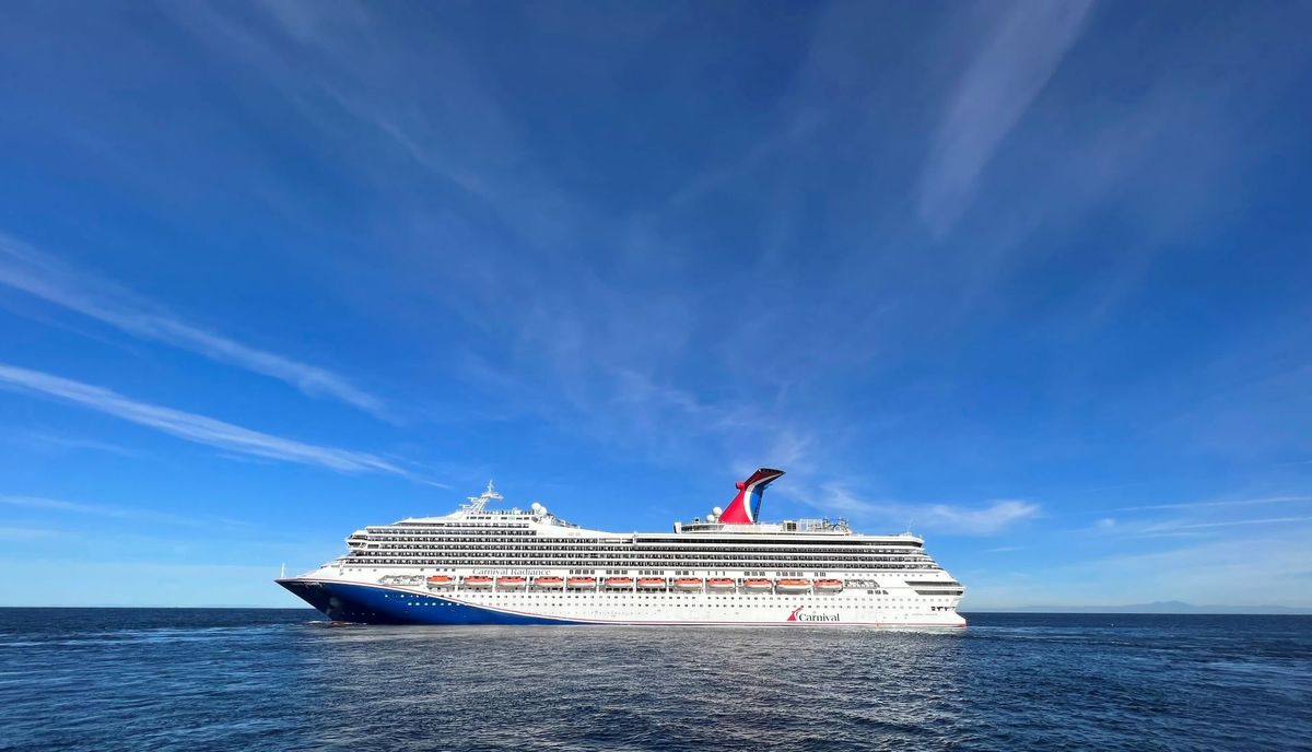 A large cruise ship sailing through calm waters under a clear blue sky. The ship features a prominent red and blue design along its hull.