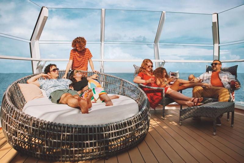 A group of six people relax and enjoy their time on a cruise ship deck. They are lounging on stylish outdoor furniture with ocean views, indicative of a vacation or leisure atmosphere.