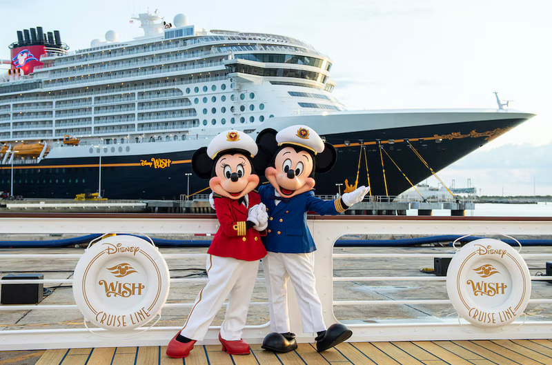 Two costumed characters resembling iconic cartoon mice pose playfully on a dock in front of a large cruise ship. The ship, emblazoned with Disney branding, is docked at a harbor, suggesting a maritime vacation setting.