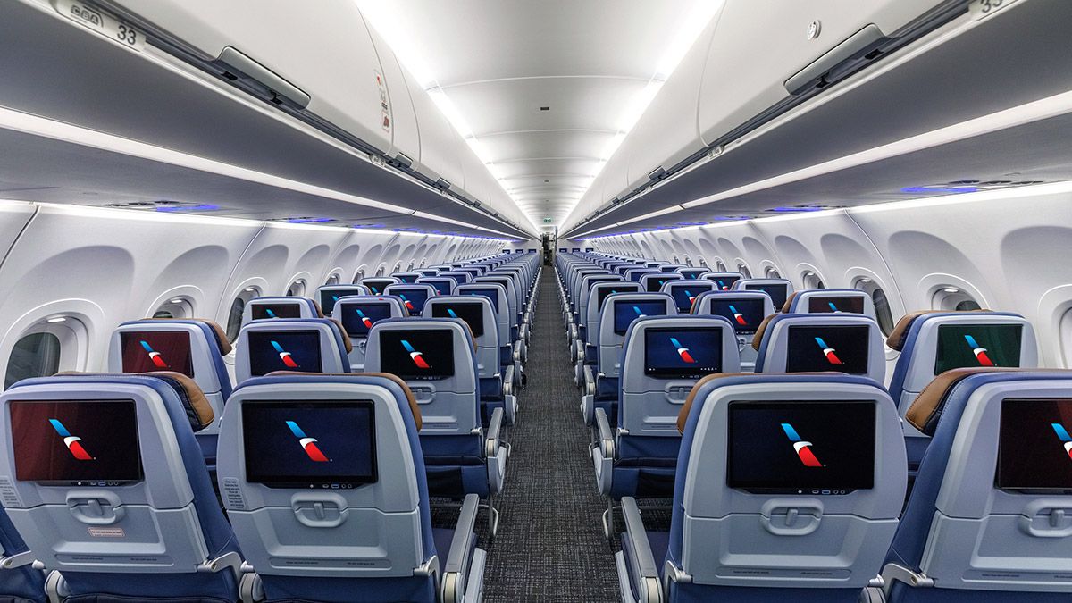 Interior view of an empty airplane cabin, showcasing rows of passenger seats. Each seat features an individual entertainment screen, and the cabin is well-lit with modern overhead lighting.