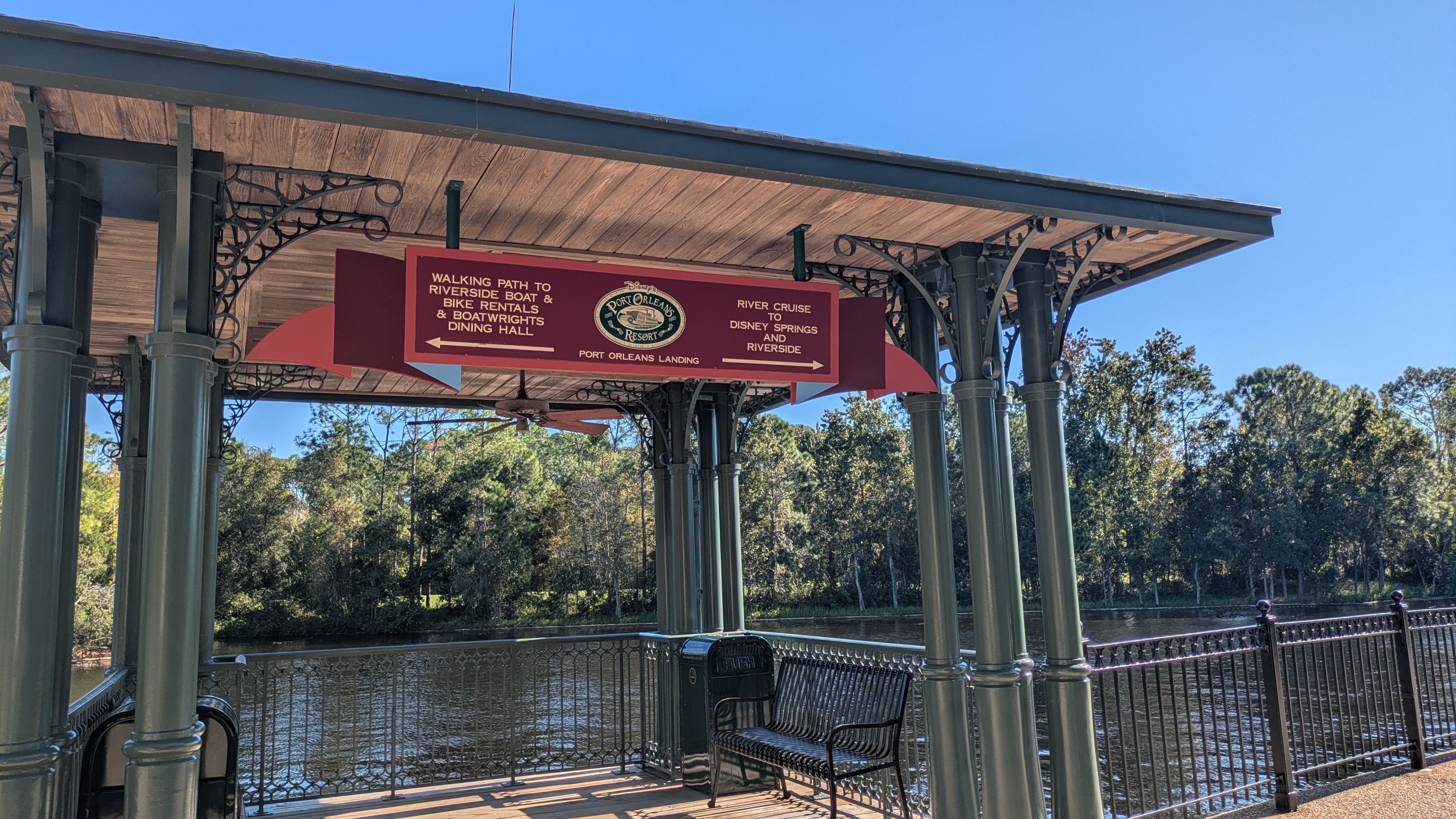 A scenic view of a waterfront dock featuring a covered structure with decorative architecture. The structure includes a sign that offers information about nearby dining options and activities such as river cruises.