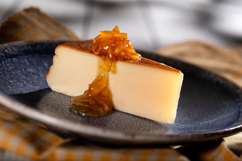 A slice of cheesecake is presented on a dark plate, topped with a shiny fruit glaze. The dessert is set against a neutral background, highlighting its creamy texture and appearance.