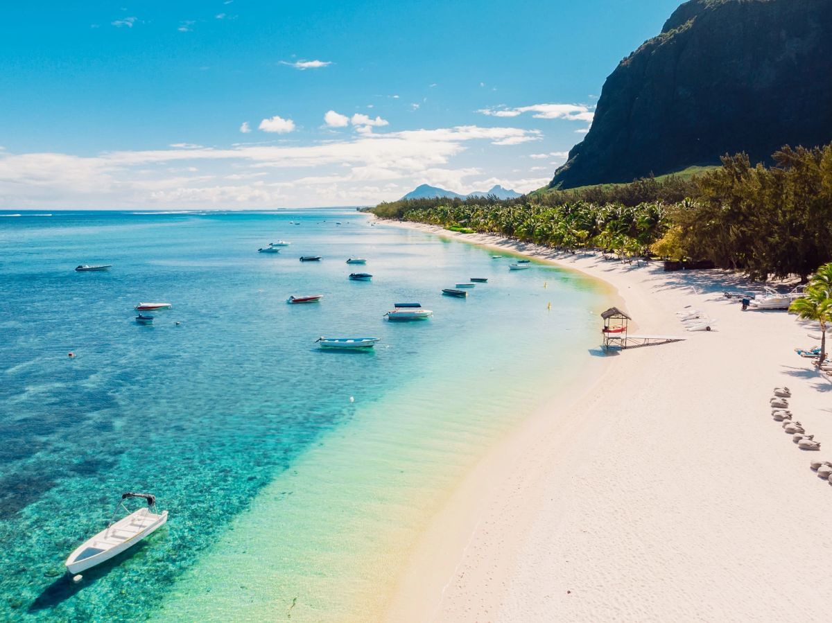 A beautiful aerial view of a calm beach with clear turquoise waters and white sand. Several boats are anchored near the shore, while lush greenery and a hilly backdrop complete the scenic landscape.