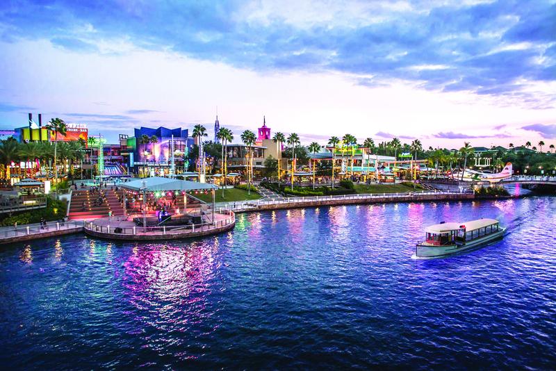 The image captures a vibrant waterfront scene featuring a shopping and entertainment area illuminated by colorful lights at dusk. A boat is seen navigating the water, while palm trees line the shore against a backdrop of a cloudy sky.