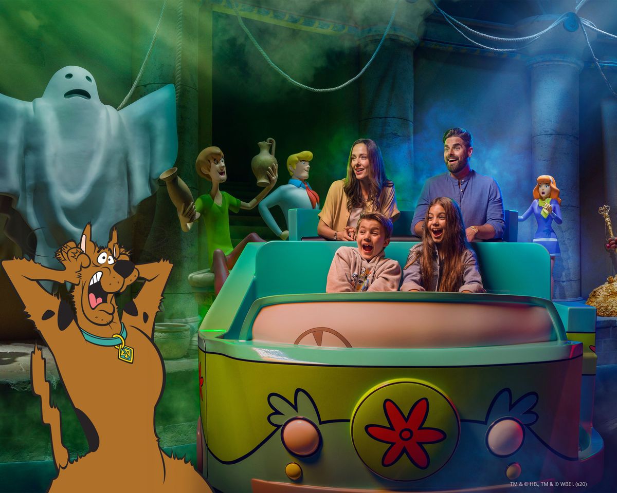 The image depicts a family enjoying a themed amusement park attraction with characters from the animated series Scooby-Doo. The scene features a ghostly figure in the background, adding to the spooky atmosphere of the ride.