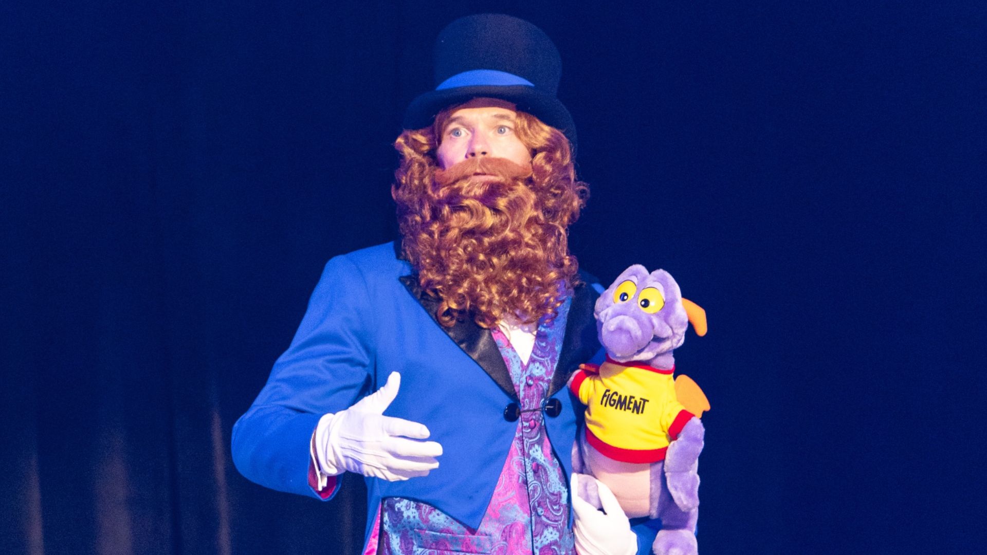 The image shows a performer dressed in a vibrant blue suit with a large black top hat. He has a long curly beard and is holding a purple puppet resembling a dragon, which is wearing a t-shirt with the word 'HOMANT' on it. The background is dark, highlighting the performer and the puppet.