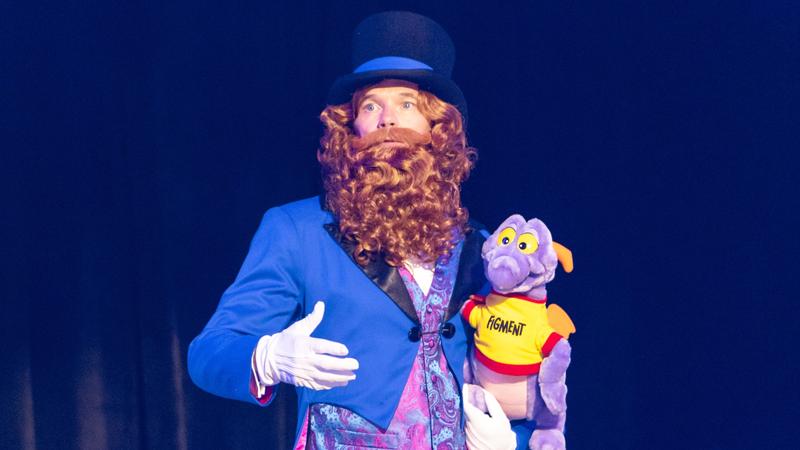 The image shows a performer dressed in a vibrant blue suit with a large black top hat. He has a long curly beard and is holding a purple puppet resembling a dragon, which is wearing a t-shirt with the word 'HOMANT' on it. The background is dark, highlighting the performer and the puppet.