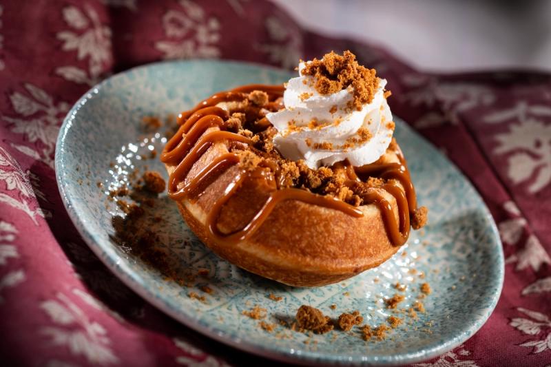 The image features a close-up of a decadent dessert, a waffle topped with caramel sauce, whipped cream, and crushed cookies, presented on a decorative blue plate. The background is a richly textured fabric that enhances the visual appeal of the dish.