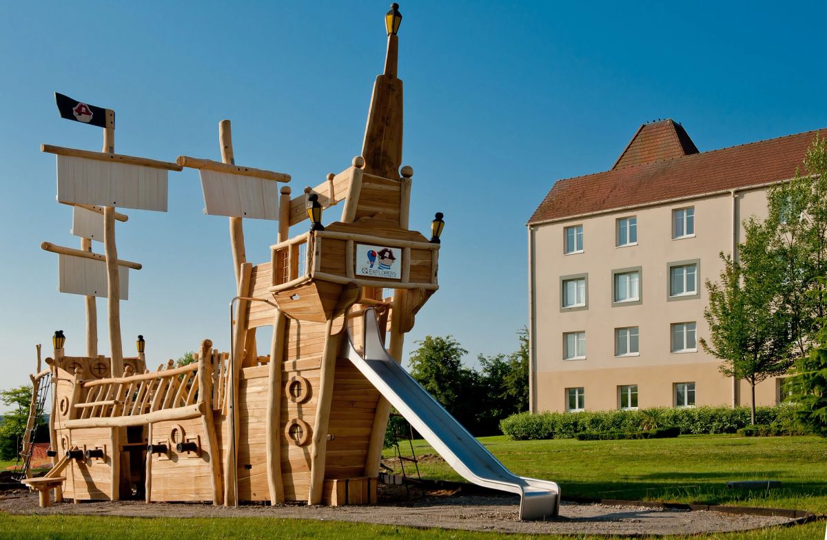 The image features a large wooden playground structure designed to resemble a pirate ship, complete with a slide and decorative sails. The playground is set in a green outdoor space beside a multi-story building.