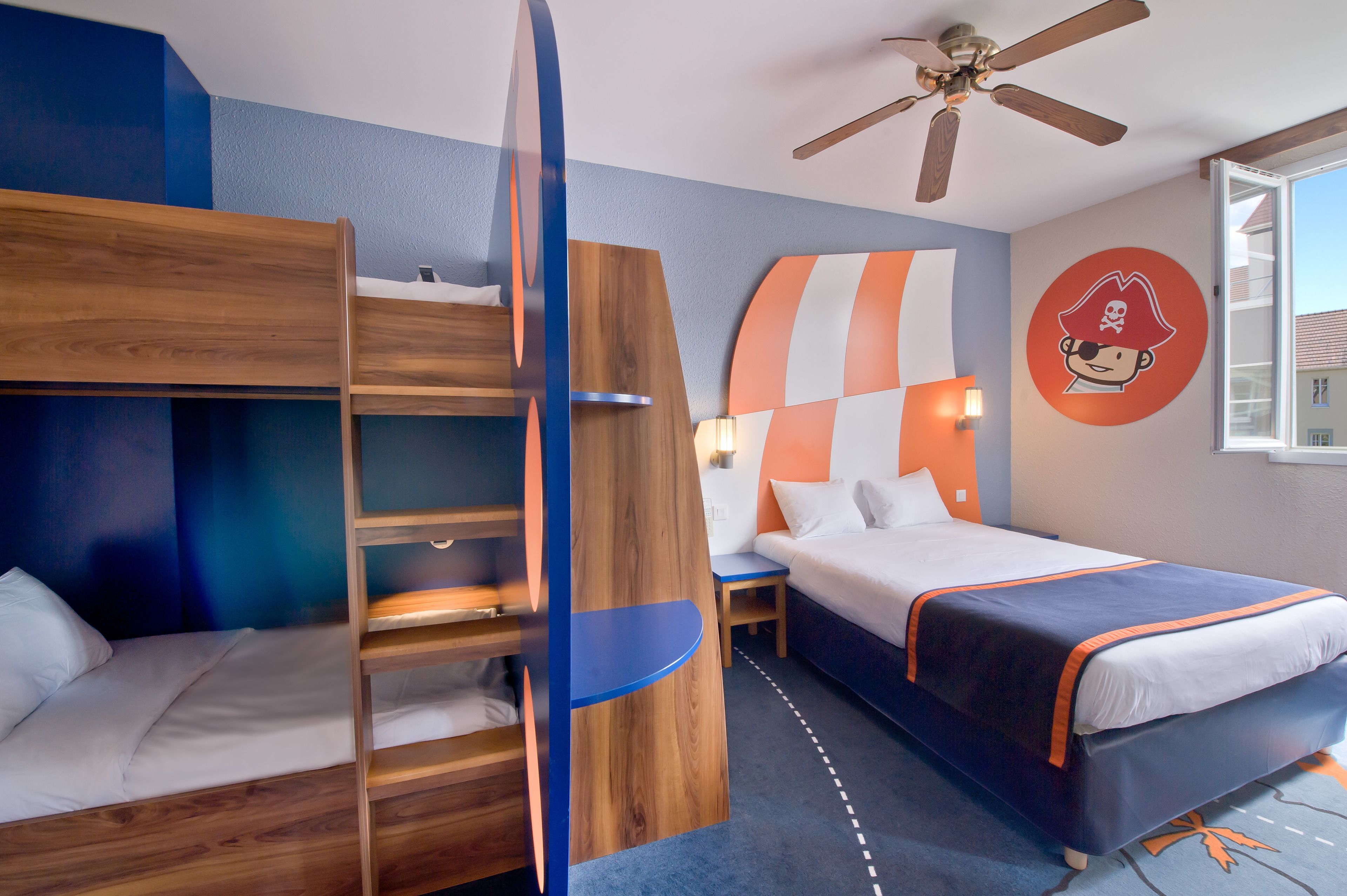 The image shows a children's themed hotel room featuring a bunk bed and a queen-sized bed. The room is decorated with a pirate motif, including a large pirate character on the wall and colorful accents.