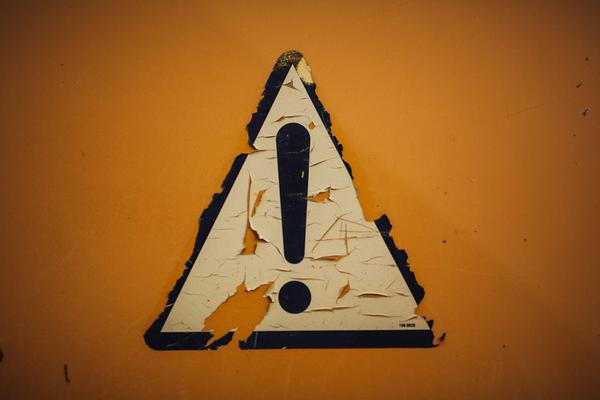 The image features a weathered yellow caution sign prominently displaying an exclamation mark. The surface of the sign is peeling and shows signs of wear, indicating it may have been exposed to the elements for some time.