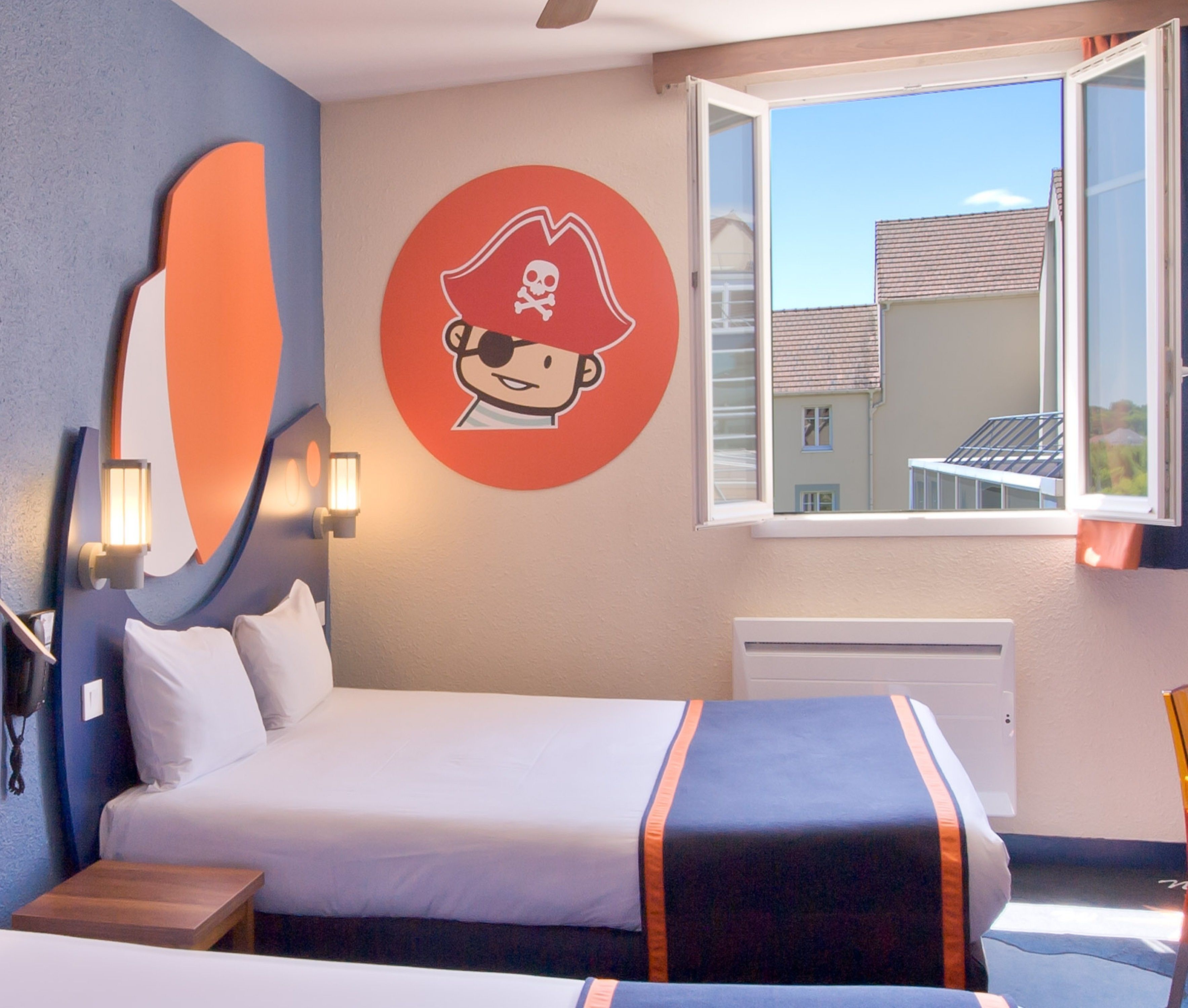The image showcases a children's hotel room featuring a playful pirate theme. It includes two beds with colorful bedding and a large mural of a pirate character on one wall, alongside a window that opens to a bright exterior view.